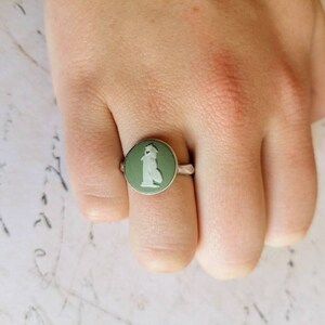 Può includere: Un anello d'argento con un cammeo verde che presenta una figura bianca. Il cammeo è incastonato in una lunetta rotonda.