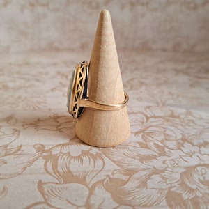 Pu&ograve; includere: Un anello d'oro con una pietra ovale bianca e nera incastonata in un design a filigrana d'oro. L'anello &egrave; esposto su un porta anelli in legno.