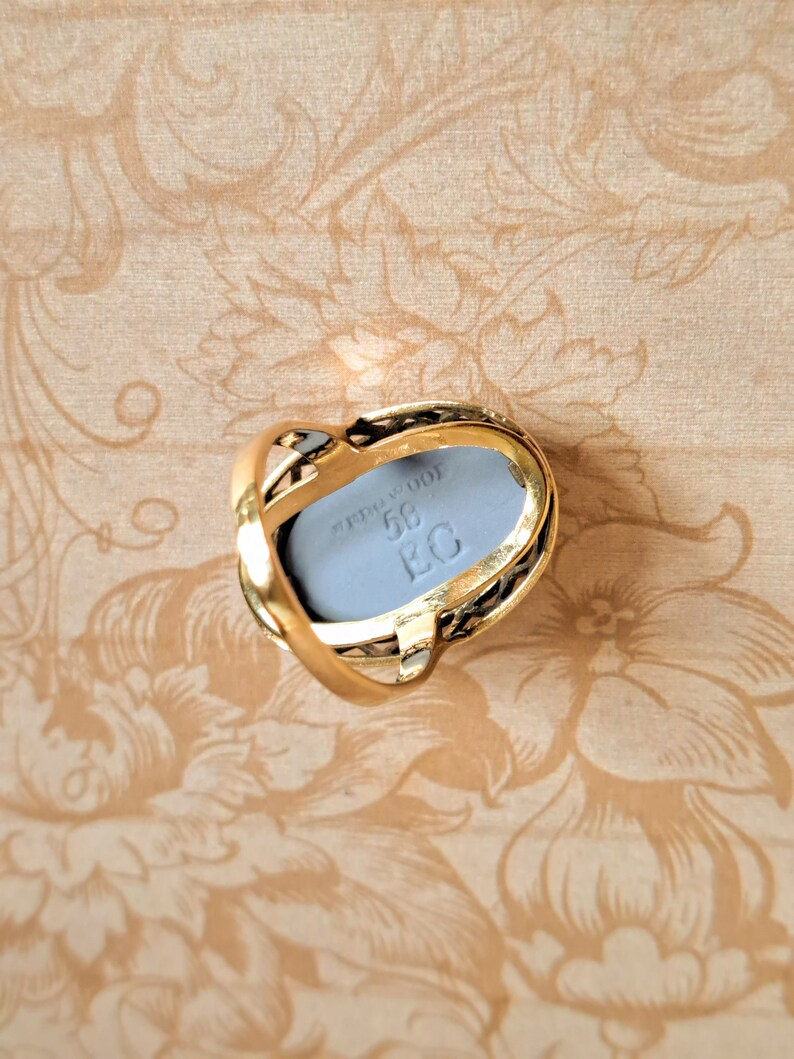Pu&ograve; includere: Un anello d'oro con una pietra ovale grigia incastonata in un design a filigrana nero e oro. La pietra ha l'iscrizione "STERLING 925 58 EG".