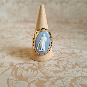 Pu&ograve; includere: Un anello con cammeo blu e bianco con una montatura in oro. Il cammeo presenta una figura bianca di una persona in piedi su uno sfondo blu.