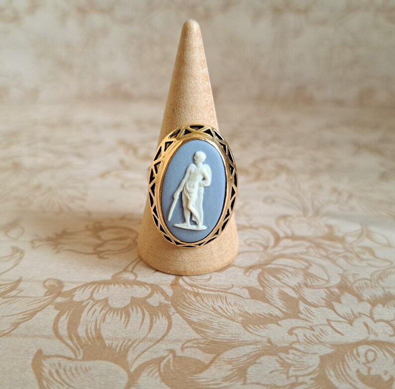 Pu&ograve; includere: Un anello con cammeo blu e bianco con una montatura in oro. Il cammeo presenta una figura bianca di una persona in piedi su uno sfondo blu.