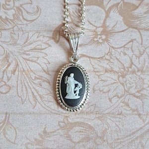 May include: A silver chain necklace with an oval pendant featuring a black cameo with a white ceramic figure of a woman holding a branch.