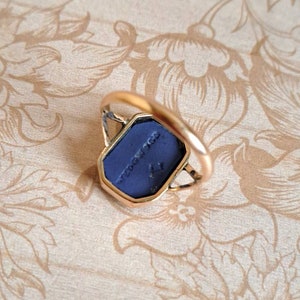 K&ouml;nnte beinhalten: Ein goldener Ring mit einem blauen Stein, der in eine geometrische Form gefasst ist. Der Stein tr&auml;gt die Inschrift "NEW YORK".