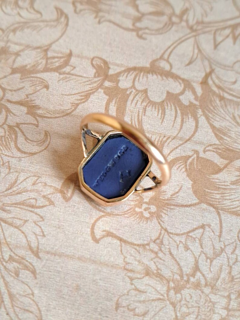 K&ouml;nnte beinhalten: Ein goldener Ring mit einem blauen Stein, der in eine geometrische Form gefasst ist. Der Stein tr&auml;gt die Inschrift "NEW YORK".
