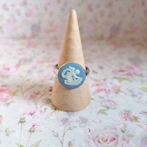 Könnte beinhalten: Ein silberner Ring mit einem blauen Kamee, der einen weißen Cherub darstellt. Der Ring wird auf einem hölzernen Ringständer präsentiert.