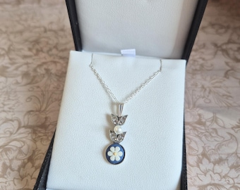 May include: A silver necklace with a pendant featuring a white flower on a blue background, set in a round frame. The necklace is presented in an open black jewellery box with a white interior. The pendant is accented with a pearl and decorative silver elements.