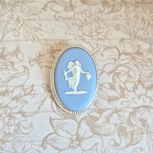 Può includere: Un broche a cammeo blu e bianco con una donna che tiene dei fiori. Il cammeo è incastonato in una cornice ovale argentata con un bordo perlato.