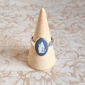 Può includere: Un anello d'argento con un cammeo nero che presenta una figura bianca di una donna che tiene una cornucopia. L'anello è esposto su un porta anelli di legno.