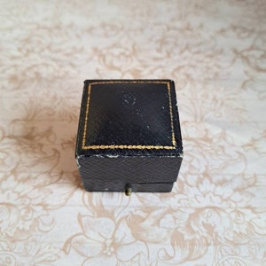 K&ouml;nnte beinhalten: Eine kleine, quadratische Ringbox in Schwarz mit einem goldenen Rand um die Kanten. Die Box ist geschlossen und steht auf einer Oberfl&auml;che mit floralem Muster.