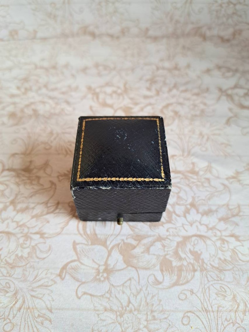 K&ouml;nnte beinhalten: Eine kleine, quadratische Ringbox in Schwarz mit einem goldenen Rand um die Kanten. Die Box ist geschlossen und steht auf einer Oberfl&auml;che mit floralem Muster.