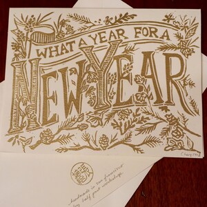 May include: A gold-colored greeting card with the words "WHAT A YEAR FOR A NEW YEAR" in a decorative font. The card is surrounded by floral and botanical illustrations. An envelope is beneath the card.