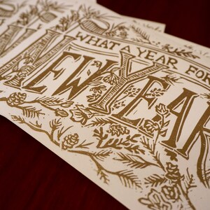 May include: A stack of cream-colored cards with gold lettering. The cards feature the phrase "WHAT A YEAR FOR A NEW YEAR" in a decorative font, surrounded by floral and botanical illustrations. The cards are arranged on a dark red surface.
