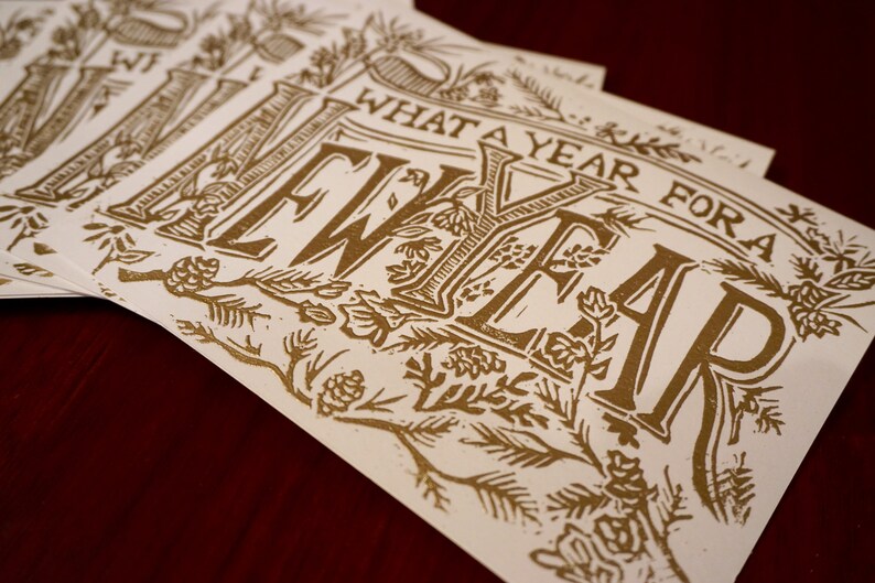 May include: A stack of cream-colored cards with gold lettering. The cards feature the phrase "WHAT A YEAR FOR A NEW YEAR" in a decorative font, surrounded by floral and botanical illustrations. The cards are arranged on a dark red surface.
