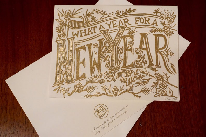 May include: A gold-colored greeting card with the words "WHAT A YEAR FOR A NEW YEAR" in a decorative font. The card is surrounded by floral and botanical illustrations. An envelope is beneath the card.