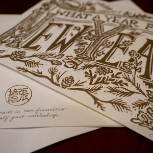 May include: A stack of white greeting cards with gold-colored text and illustrations. The cards feature the phrase "WHAT A YEAR FOR A NEW YEAR" in a decorative font. The bottom card has a stamp and the text "handmade in san francisco by half past workshop."