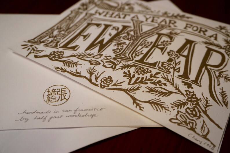 May include: A stack of white greeting cards with gold-colored text and illustrations. The cards feature the phrase "WHAT A YEAR FOR A NEW YEAR" in a decorative font. The bottom card has a stamp and the text "handmade in san francisco by half past workshop."