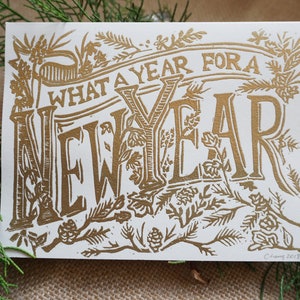May include: A rectangular greeting card with the words "WHAT A YEAR FOR A NEW YEAR" in gold lettering. The card is decorated with botanical illustrations and is set against a backdrop of greenery.