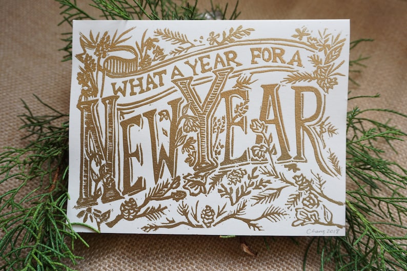 May include: A rectangular greeting card with the words "WHAT A YEAR FOR A NEW YEAR" in gold lettering. The card is decorated with botanical illustrations and is set against a backdrop of greenery.