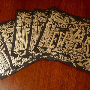 May include: A stack of black cards with gold foil lettering and floral illustrations. The cards read "WHAT A YEAR FOR A NEW YEAR" in an elegant, vintage font. The cards are arranged on a dark wood surface.