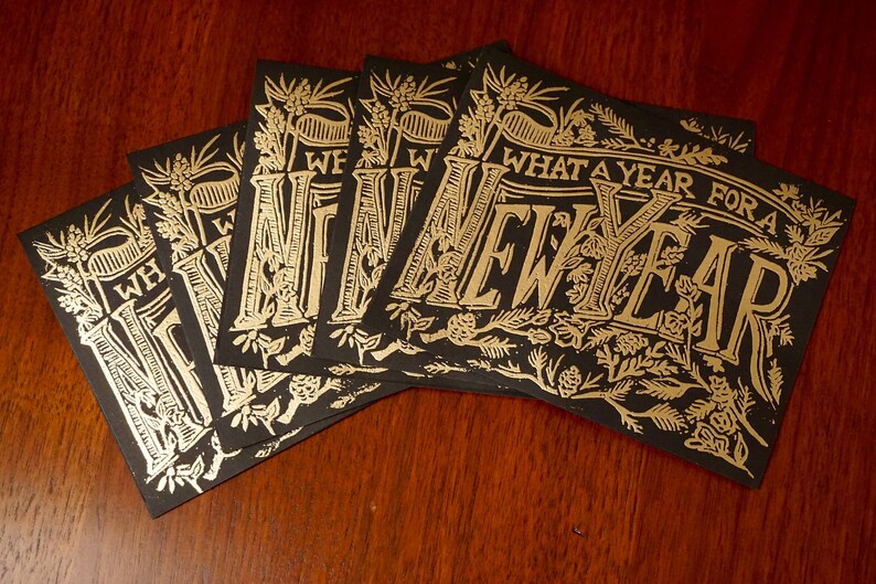 May include: A stack of black cards with gold foil lettering and floral illustrations. The cards read "WHAT A YEAR FOR A NEW YEAR" in an elegant, vintage font. The cards are arranged on a dark wood surface.