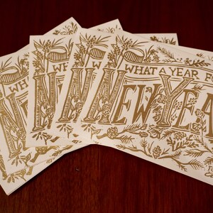 May include: A collection of cream-colored cards with gold lettering and botanical illustrations. The cards display the phrase "WHAT A YEAR FOR A NEW YEAR" in a decorative font. The cards are arranged on a dark wood surface.