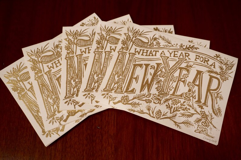 May include: A collection of cream-colored cards with gold lettering and botanical illustrations. The cards display the phrase "WHAT A YEAR FOR A NEW YEAR" in a decorative font. The cards are arranged on a dark wood surface.