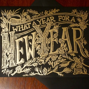 May include: A black greeting card with gold foil lettering and floral embellishments. The text reads "WHAT A YEAR FOR A NEW YEAR". The card is placed on a black envelope, suitable for sending holiday greetings.