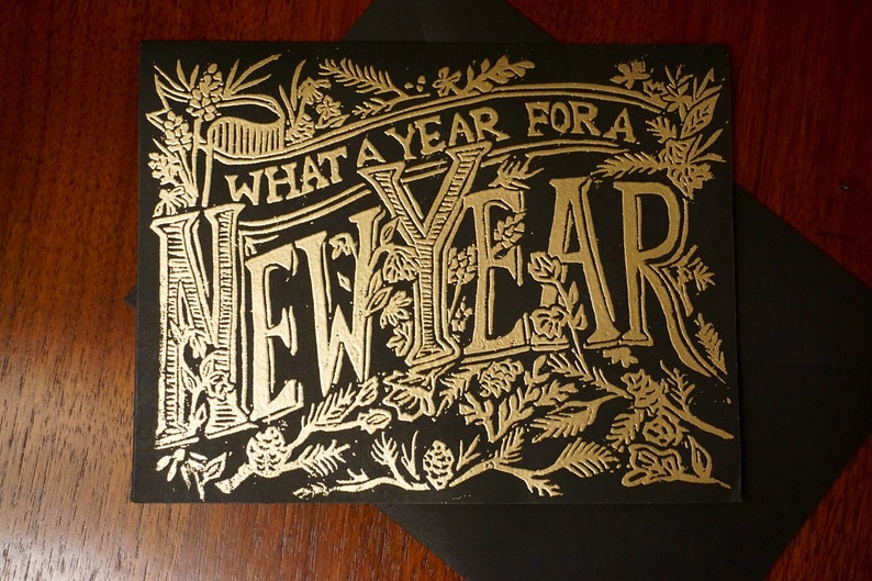 May include: A black greeting card with gold foil lettering and floral embellishments. The text reads "WHAT A YEAR FOR A NEW YEAR". The card is placed on a black envelope, suitable for sending holiday greetings.