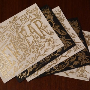 May include: A collection of square cards with gold foil lettering and floral designs. The cards feature the text "WHAT A YEAR FOR A" and are arranged on a dark wood surface. Some cards are black with gold designs.