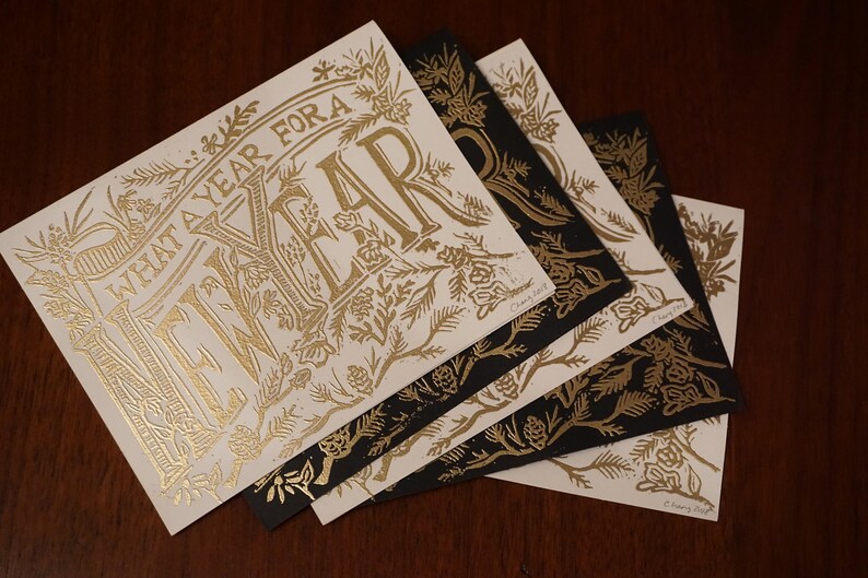 May include: A collection of square cards with gold foil lettering and floral designs. The cards feature the text "WHAT A YEAR FOR A" and are arranged on a dark wood surface. Some cards are black with gold designs.