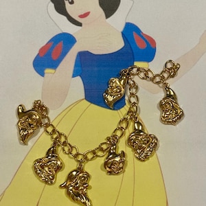 May include: A gold charm bracelet with six detailed charms of the seven dwarfs from Snow White. The bracelet is displayed in front of a Snow White illustration. The charms are connected by a gold chain.