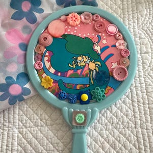 May include: A light blue hand mirror with a round, pink and blue patterned surface. The surface is decorated with colorful buttons in various shapes and sizes.