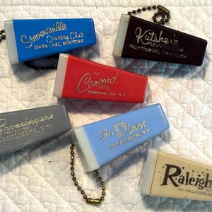 Puede incluir: Una colección de llaveros de hotel vintage en varios colores, incluyendo azul, rojo, gris y beige. Cada llavero presenta el nombre y la ubicación de un hotel en letras doradas. Los llaveros están unidos a pequeñas cadenas de bolas de metal.