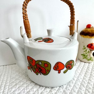 Peut inclure: Une théière en céramique blanche ornée d'un motif de champignons colorés dans des tons orange et vert olive. La théière possède un bec verseur incurvé, un couvercle assorti et une anse haute arquée en osier brun naturel, avec un petit pot en forme de champignon.