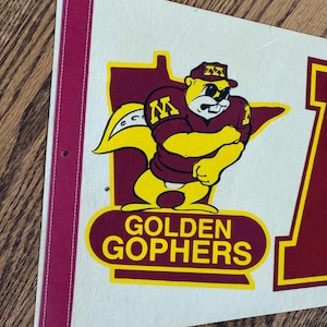 Könnte beinhalten: Eine cremefarbene Wimpel mit einem weinroten Rand und Nähten. Der Wimpel zeigt einen gelben und weinroten Cartoon-Gopher, der eine Football-Uniform trägt, und die Worte "GOLDEN GOPHERS". Der Gopher ist vor einer weinroten Umrisslinie des Bundesstaates Minnesota platziert.