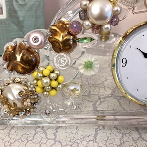 Puede incluir: Un reloj de cristal transparente con un borde dorado y una esfera blanca. El reloj está decorado con varias cuentas, pedrería y flores. La esfera del reloj dice "WALTHER" y tiene agujas negras.