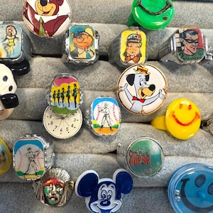 May include: A collection of vintage rings featuring cartoon characters, a smiley face, and other designs. The rings are made of various materials and have different colored bands. Some rings have a silver band.