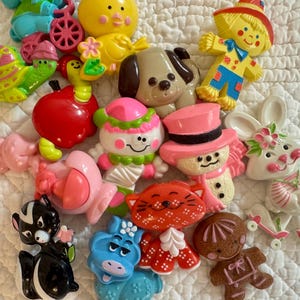 Puede incluir: Una colección de broches de plástico de colores que incluye un pollito amarillo, un espantapájaros, un perro marrón, una manzana roja con un gusano, un caracol verde, un muñeco de nieve rosa, un muñeco de nieve con sombrero rosa, un conejo y un mofeta.