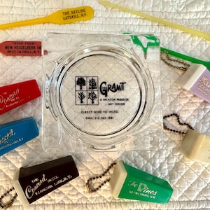 以下が含まれることがあります： 「The Granit A Vacation Paradise Any Season Direct Wire to Hotel Dial 212-563-1881」と書かれた透明な正方形のガラス製灰皿。周囲には、ニューヨーク州キャッツキルの文字が印刷されたヴィンテージのプラスチック製ホテルキーホルダーやドリンクマドラーが配置されています。