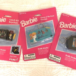 Può includere: Tre carte di imballaggio rosa e bianche per portachiavi Barbie. Ogni carta presenta un portachiavi diverso: una scatola per il pranzo, una scatola per cappelli e un portachiavi Queen of the Prom. I portachiavi sono tutti argentati e neri con il marchio Barbie.