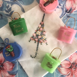 Op de afbeelding: Vier plastic regenkapjes-etuis in groen, roze, lichtroze en blauw, elk met een goudkleurig hengsel en de tekst "Rain Bonnet" in gouden letters. De etuis zijn gerangschikt rond een witte zakdoek met een geborduurd bloemenparaplu-motief.