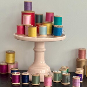 May include: A collection of colorful spools of thread, arranged on a pink cake stand and a dark surface. The spools are in various colors, including pink, yellow, green, blue, and purple. The thread spools are made of wood and are of different sizes.
