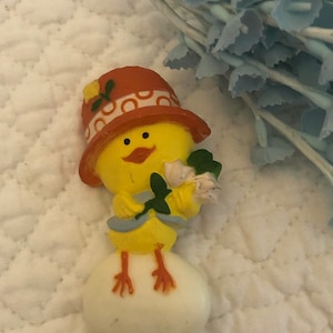 May include: A yellow chick wearing an orange and white hat with a green leaf and white flowers in its hand, sitting on a white egg.