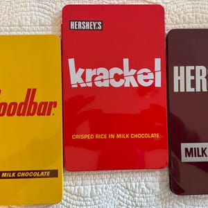 May include: Three rectangular metal tins with different Hershey's chocolate bar designs. The first tin is yellow with the text "HERSHEY'S mr. Goodbar PEANUTS IN MILK CHOCOLATE". The second tin is red with the text "HERSHEY'S krackel CRISPED RICE IN MILK CHOCOLATE". The third tin is maroon with the text "HERSHEY'S MILK CHOCOLATE".