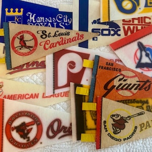 Peut inclure: Une collection de fanions de sport en feutre vintage présentant divers logos et noms d'équipes de la Major League Baseball, dont les St. Louis Cardinals, San Francisco Giants, San Diego Padres, Baltimore Orioles et Kansas City Royals, disposés en couches.