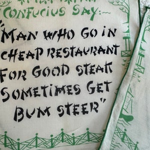 May include: Two white fabric napkins with green borders and a black and white graphic print. The text on the napkins reads "Confucius Say: Man who go in cheap restaurant for good steak sometimes get bum steer."