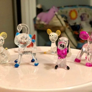 Puede incluir: Cinco pequeñas figuritas de animales de vidrio transparente en un plato blanco. Las figuritas son un cisne, un ratón, un gato, un perro y un elefante. El cisne tiene detalles azules y rojos, el ratón tiene orejas amarillas, el gato tiene detalles azules y rojos, el perro tiene detalles rosas y el elefante tiene detalles rosas.