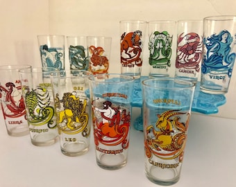 May include: Set of 12 clear glass tumblers with zodiac signs printed on them. Each glass features a different zodiac sign with its corresponding symbol and name. The signs include Libra, Scorpio, Leo, Sagittarius, Capricorn, Taurus, Gemini, Cancer, Virgo, Aries, Pisces, and Aquarius.