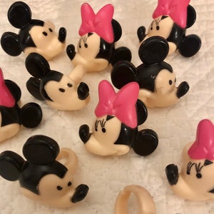 May include: A set of ten plastic Mickey Mouse and Minnie Mouse character rings. The rings are in black, white, and pink. The characters are smiling and have large ears.
