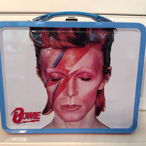 May include: A blue metal lunchbox with a picture of David Bowie on the front. The image is from the album cover for "Aladdin Sane". The album title "BOWIE" is printed in white letters on the bottom left corner of the lunchbox.